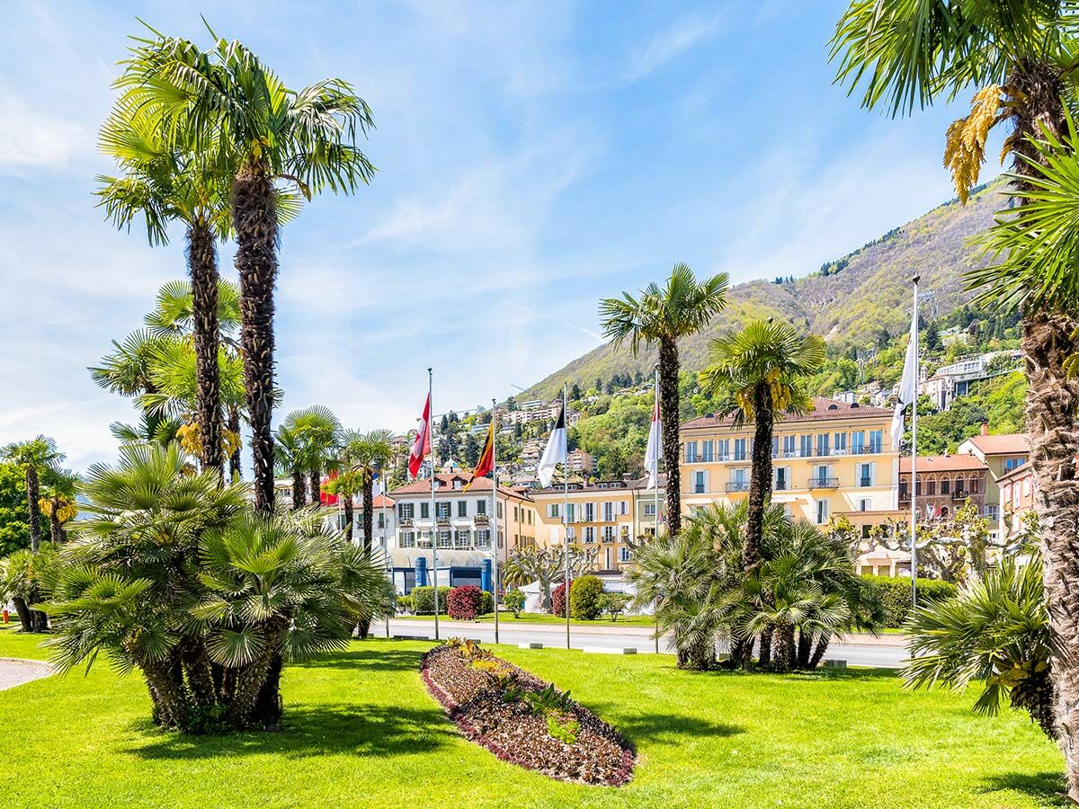 Panoramablick auf eine sonnige Palmenpromenade an einem See, mit historischen Gebäuden, Bergen und strahlend blauem Himmel.