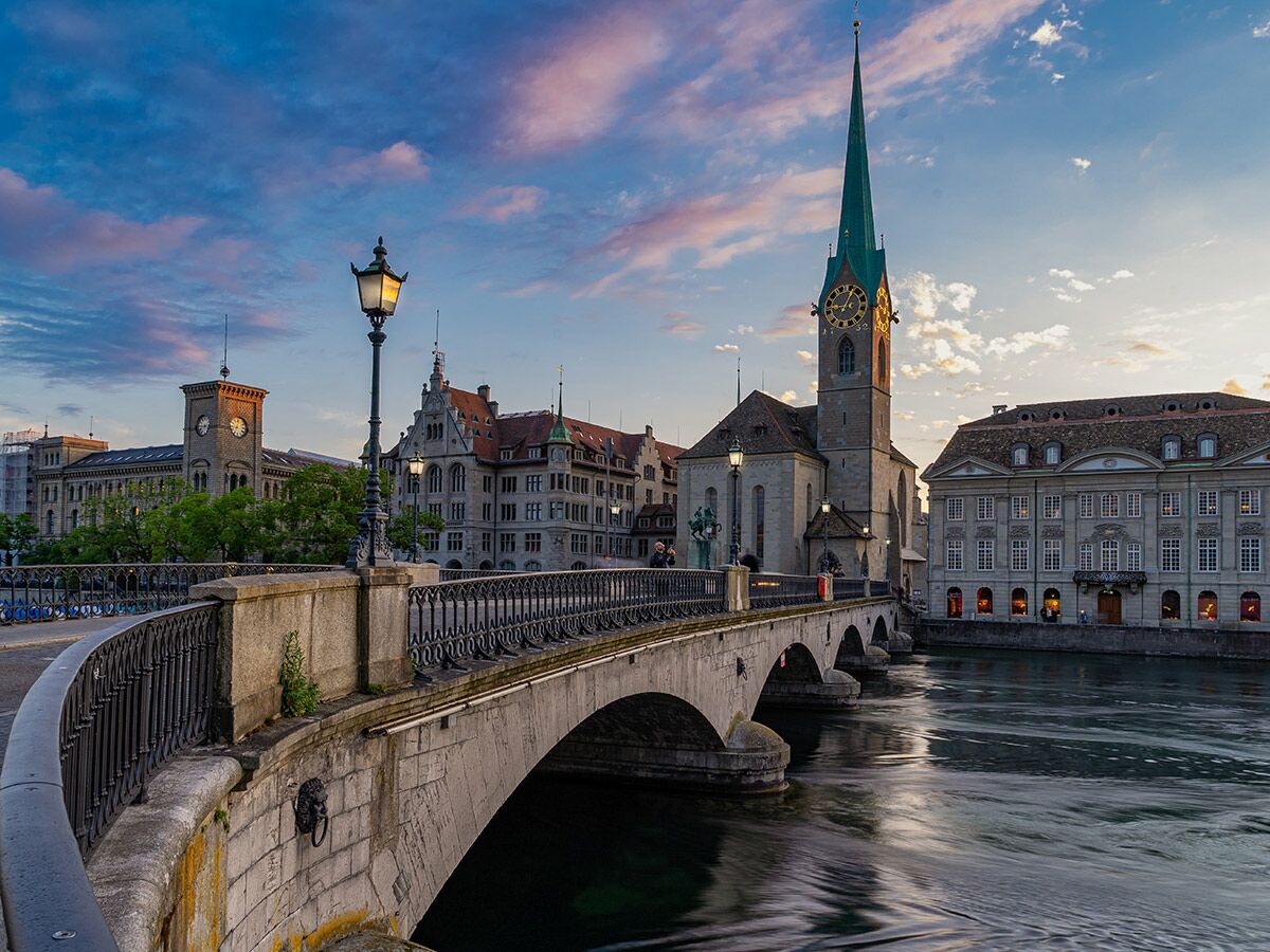 Historische Münsterbrücke über die Limmat in Zürich bei Sonnenuntergang, mit Fraumünsterkirche und Altstadtgebäuden.