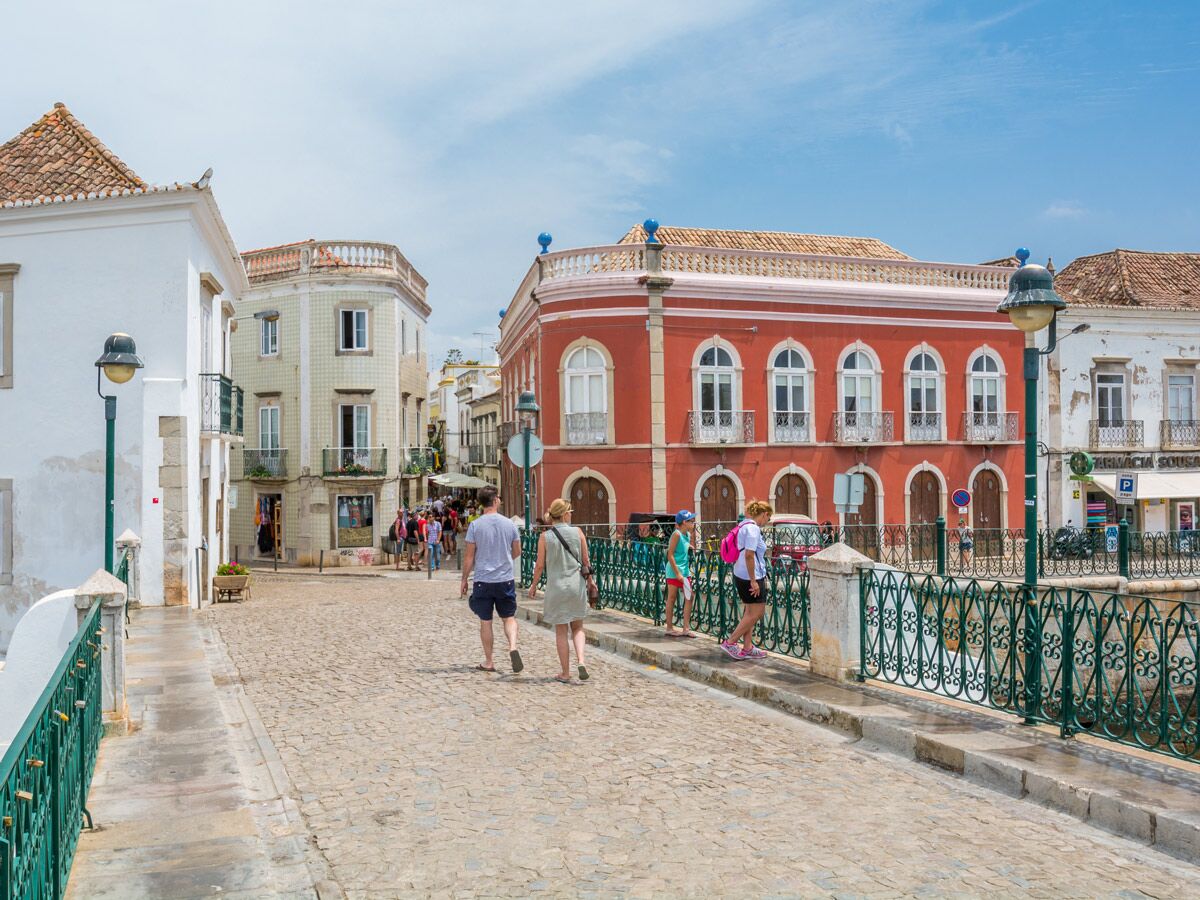 Menschen gehen auf einer Kopfsteinpflasterstraße entlang historischer Gebäude und einer grünen Brücke an einem sonnigen Tag in Portugal.