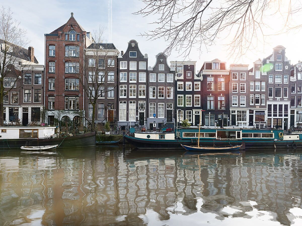 Historische Häuser und Hausboote entlang eines Amsterdamer Kanals mit Gebäudereflexionen im Wasser an einem sonnigen Tag.