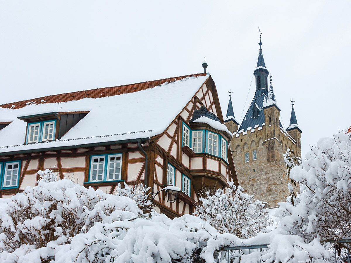 Verschneite Fachwerkhäuser mit blau umrahmten Fenstern und historischer Turm unter bewölktem Winterhimmel in Deutschland.