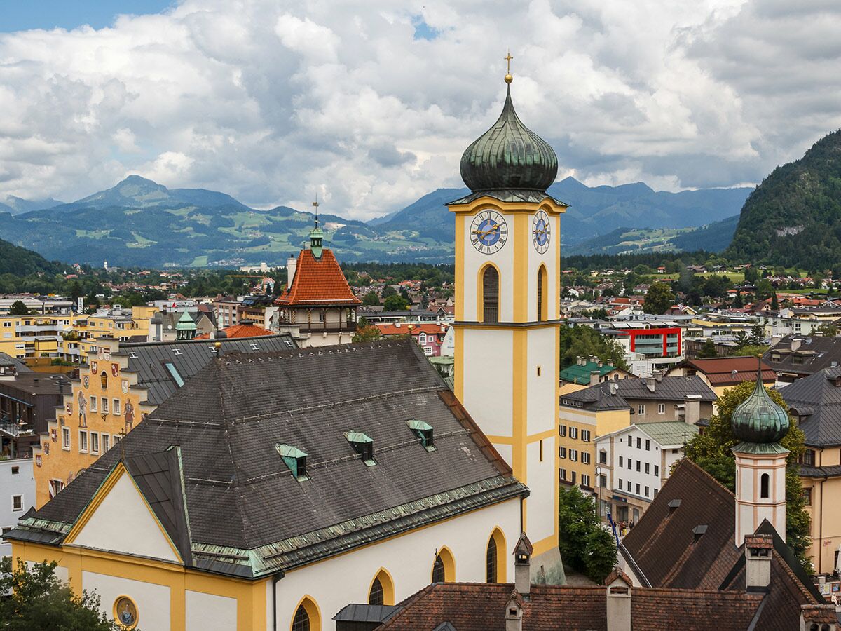 Panoramablick auf eine alpine Stadt mit prominentem Kirchturm, Zwiebelkuppel, Dächern und grünen Bergen unter Wolkenhimmel.