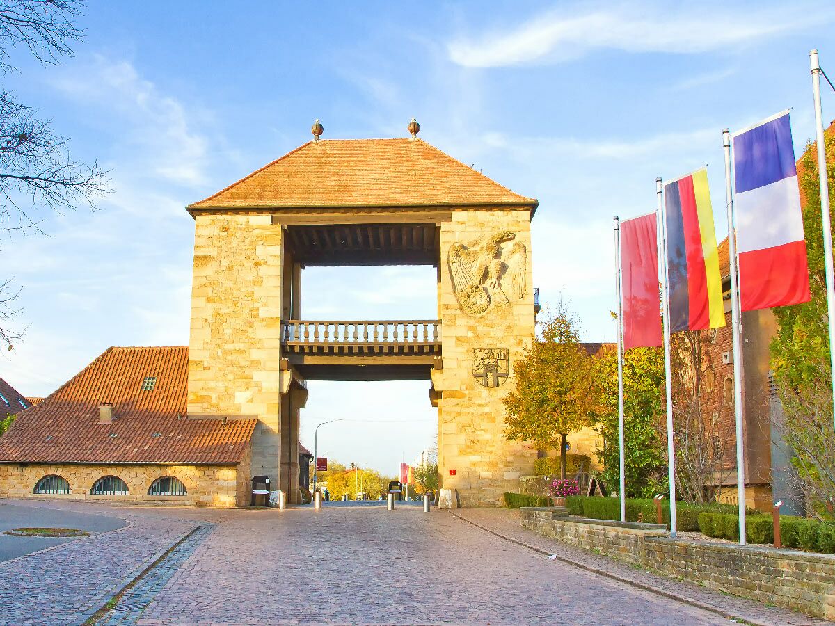 Altes Steintor mit Holzbrücke und Ziegeldach, davor wehen rote, deutsche und französische Flaggen unter blauem Himmel.