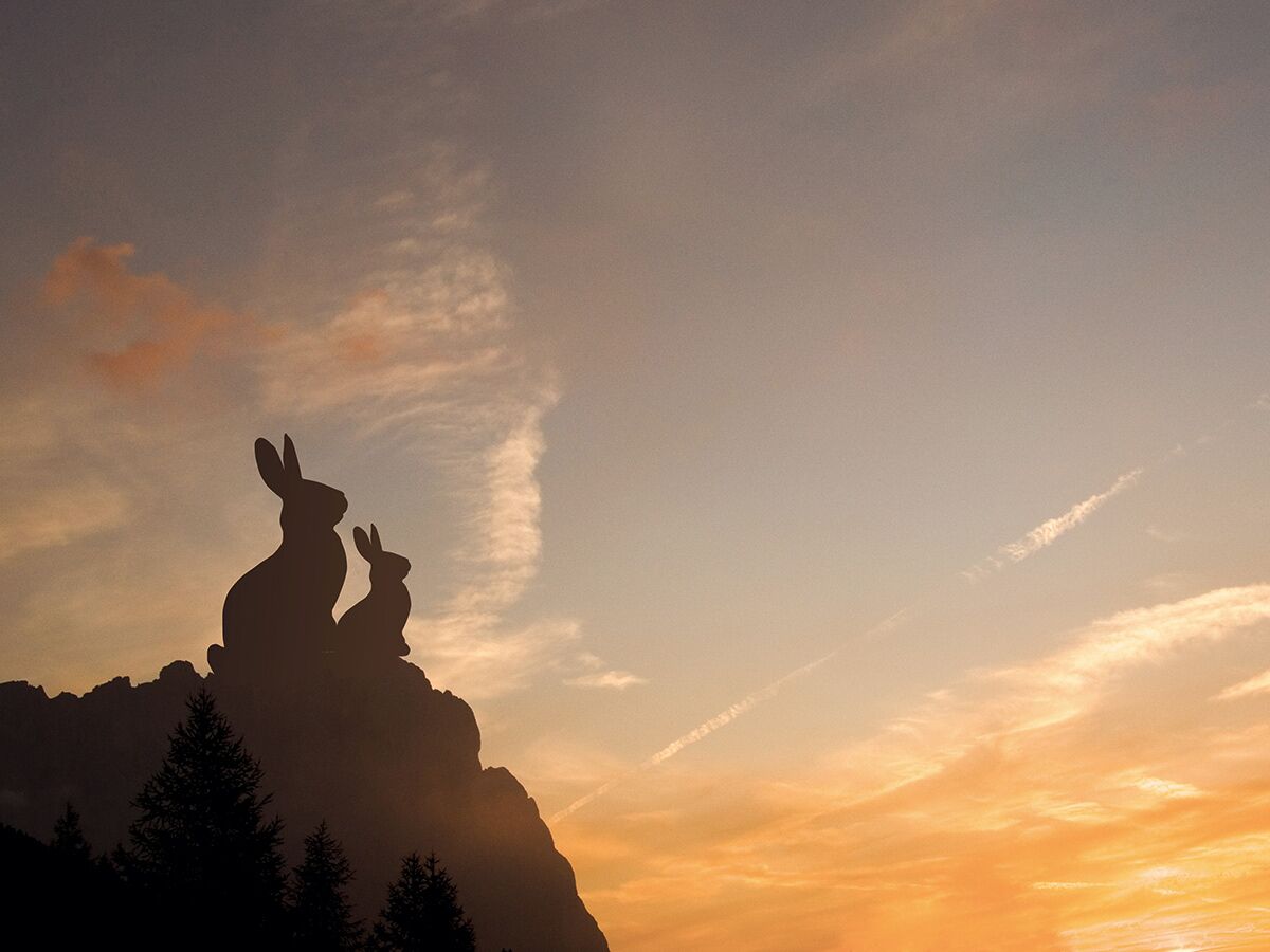 Silhouetten von zwei Hasen auf einem Berggipfel bei Sonnenuntergang, mit orange-blauem Himmel und Wolken.