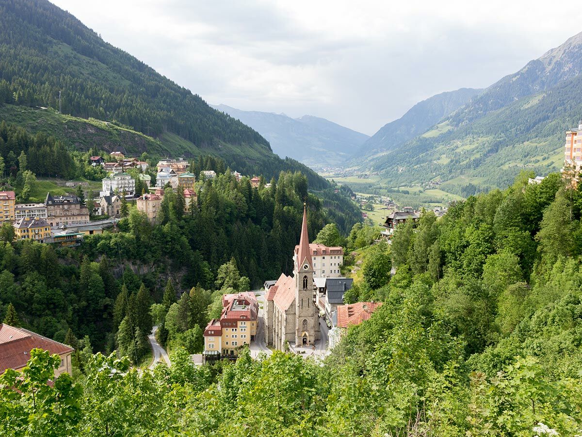 Panoramablick auf ein Bergdorf mit einer markanten Kirche, umgeben von grünen Wäldern und einem weiten Tal.