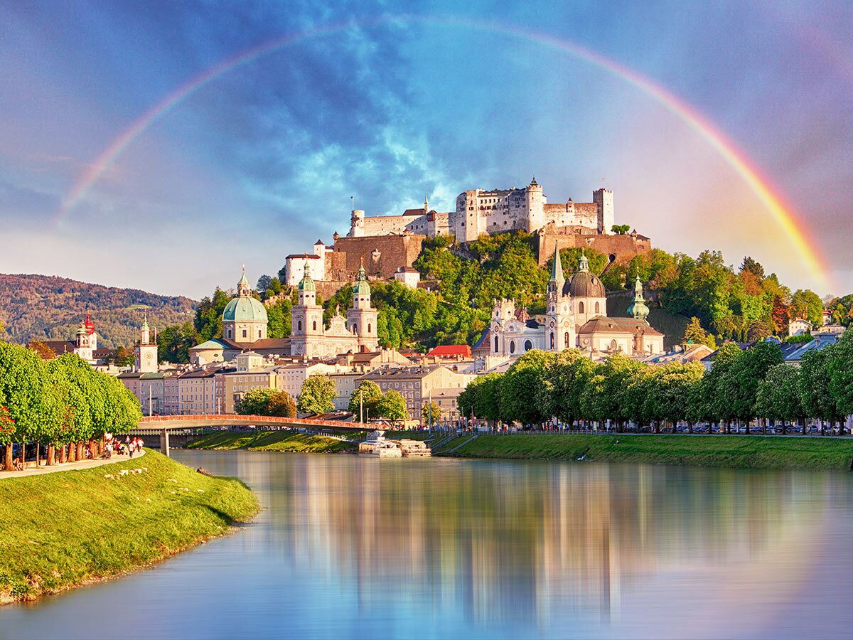 Panoramablick auf Salzburg mit der Festung Hohensalzburg, historischen Gebäuden, dem Fluss Salzach und einem Regenbogen am Himmel.