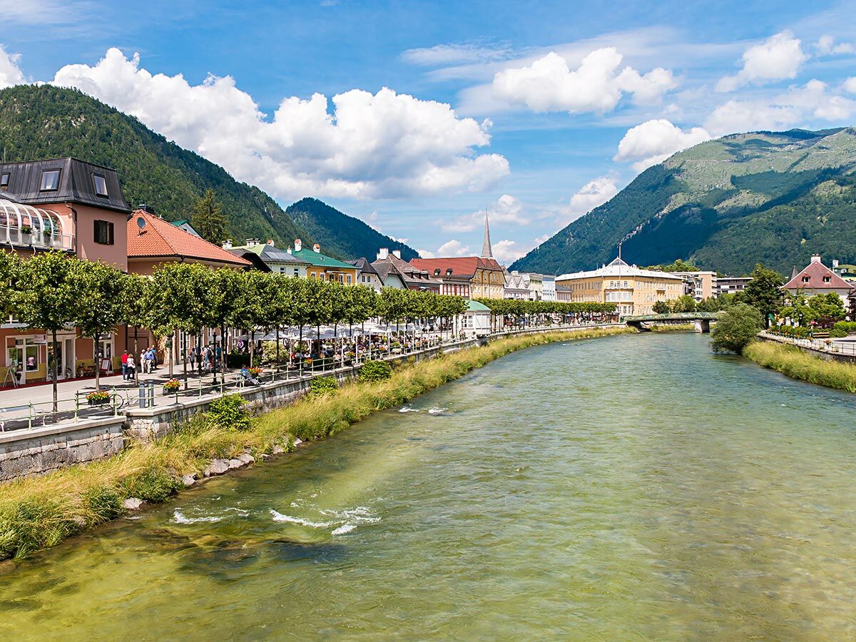 Malerische Alpenstadt am Fluss mit belebter Promenade, Cafés, historischen Gebäuden und grünen Bergen unter blauem Himmel.