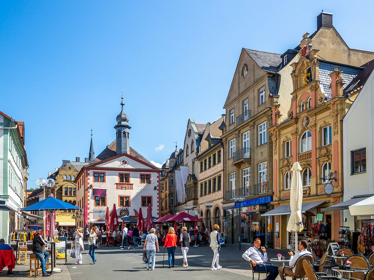 Belebter, sonnenbeschienener Marktplatz einer historischen deutschen Stadt mit Menschen, die draußen essen und einkaufen, umgeben von bunten Gebäuden und einem Rathausturm.