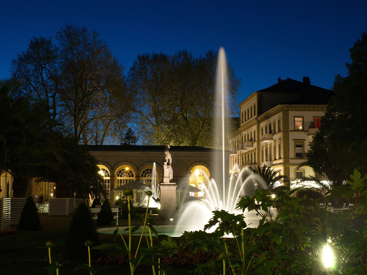 Abendaufnahme eines Parks mit beleuchtetem Springbrunnen, historischem Gebäude, Bäumen und einer Skulptur unter blauem Himmel bei Dämmerung.
