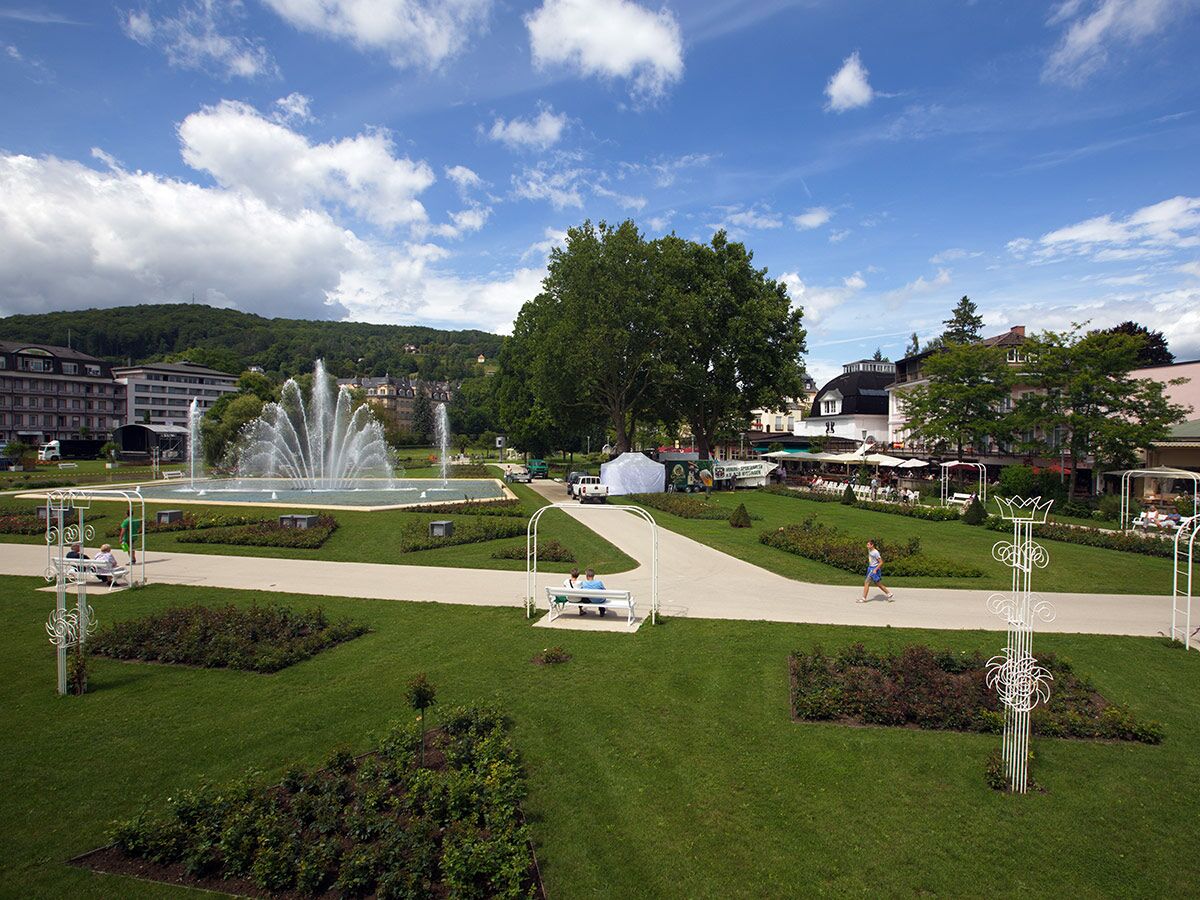 Panoramablick auf einen weitläufigen Park mit großem Springbrunnen, grünen Rasenflächen, Spazierwegen und Gebäuden im Hintergrund.