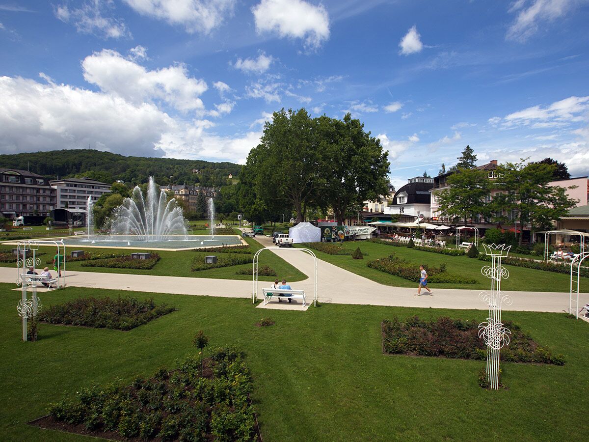 Panoramablick auf einen Stadtpark mit großem Springbrunnen, Grünflächen, Spazierwegen und Menschen an einem sonnigen Tag.