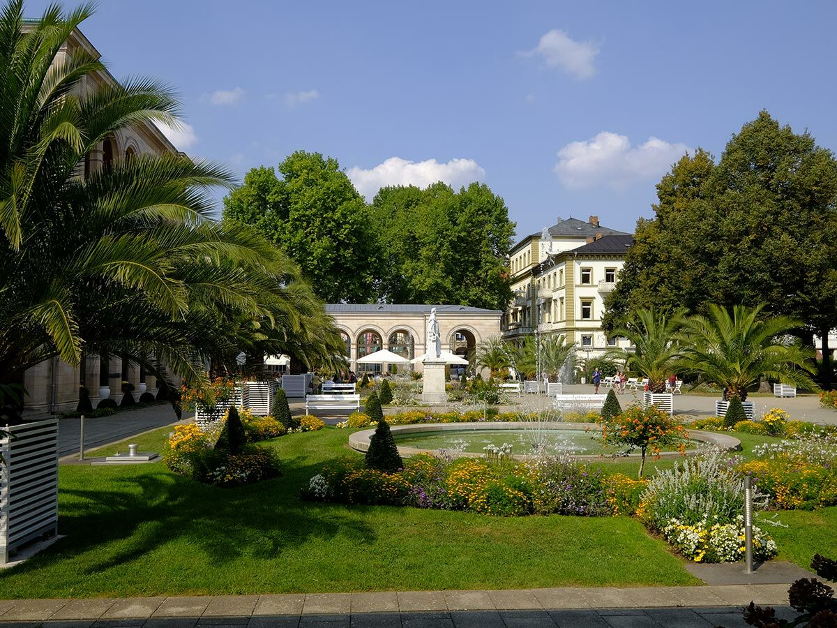 Sonniger Kurpark in Bad Kissingen mit sprudelndem Brunnen, blühenden Blumenbeeten, Palmen und historischen Gebäuden im Hintergrund.