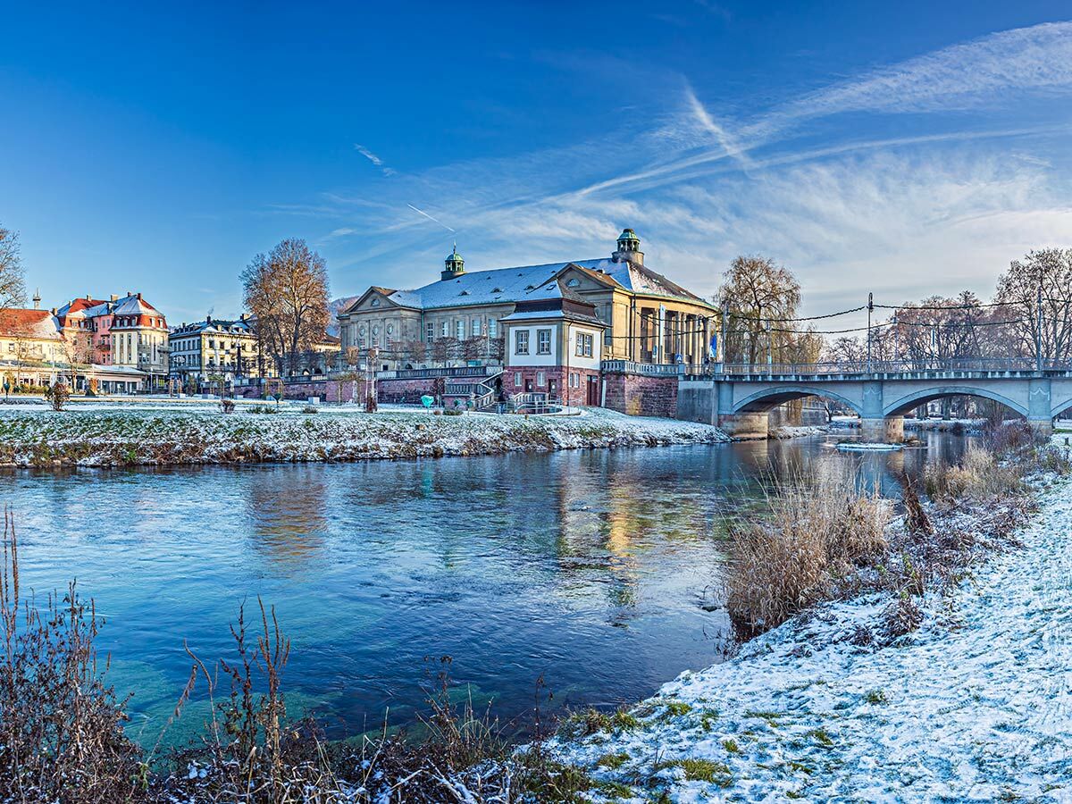 Winterlandschaft Baden-Baden: Fluss, verschneite Ufer, historische Gebäude, Bogenbrücke, blauer Himmel.