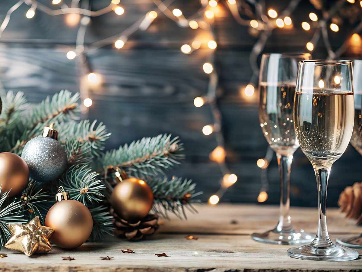 Zwei Sektgläser neben Tannenbaumzweigen mit goldenen und silbernen Weihnachtskugeln, Stern, Zapfen vor funkelnder Lichterkette auf Holztisch.