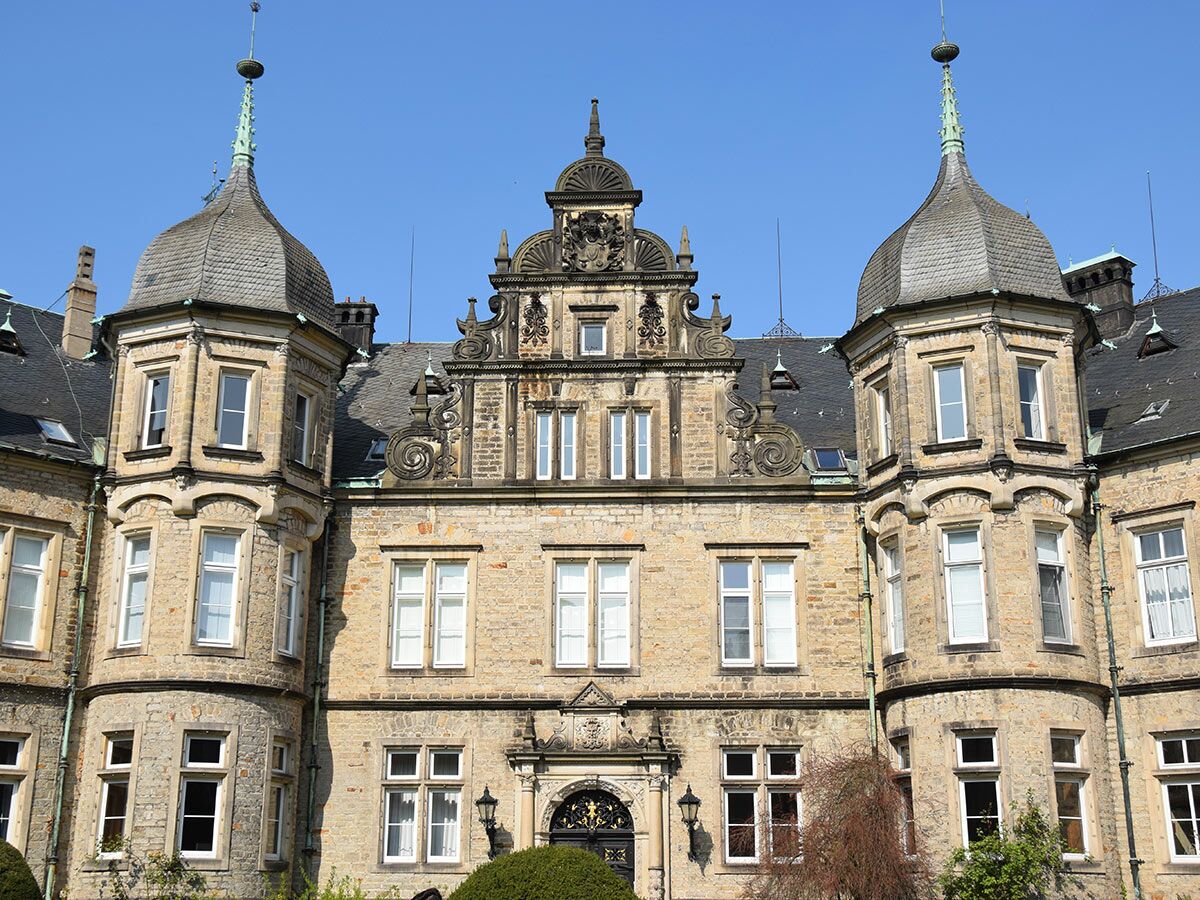 Frontansicht eines historischen Sandsteinschlossgebäudes mit zwei markanten Türmen, vielen Fenstern und detailreichem Mittelgiebel unter blauem Himmel.