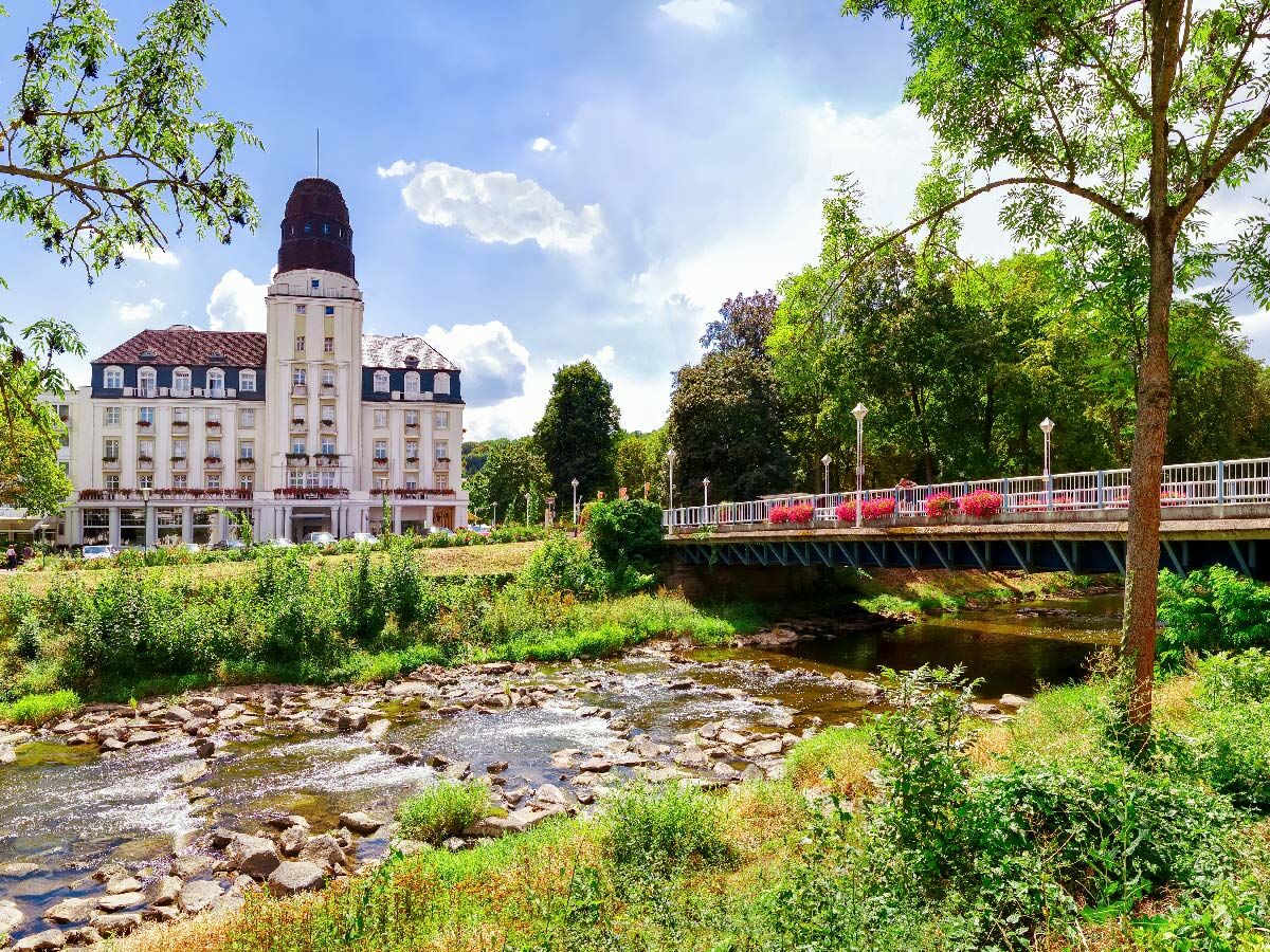 Weißes historisches Kurhotel mit markantem Turm an einem Fluss mit Felsen. Grüne Parklandschaft und eine Brücke mit roten Blumen.
