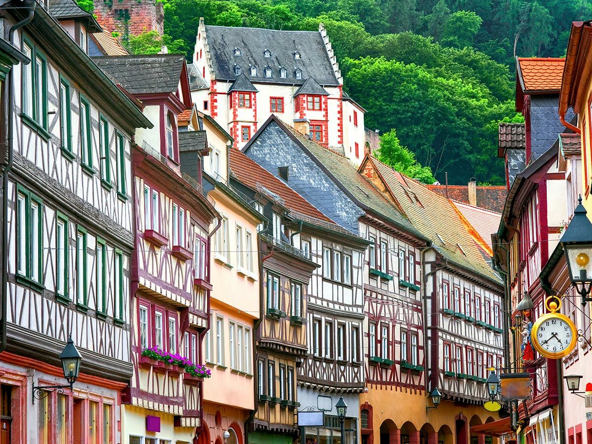 Historische deutsche Altstadtgasse mit bunten Fachwerkhäusern, Burg im Hintergrund und einer dekorativen Uhr an der Wand.