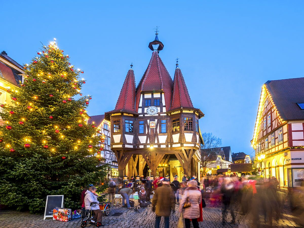 Festlicher Weihnachtsmarkt in Deutschland mit beleuchtetem Christbaum und historischem Fachwerkgebäude bei Dämmerung.