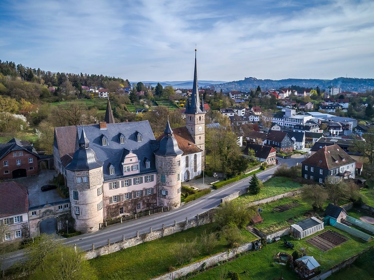 Luftbild eines historischen Schlosses und einer Kirche mit hohem Turm in einer grünen Hügellandschaft mit Kleinstadt.