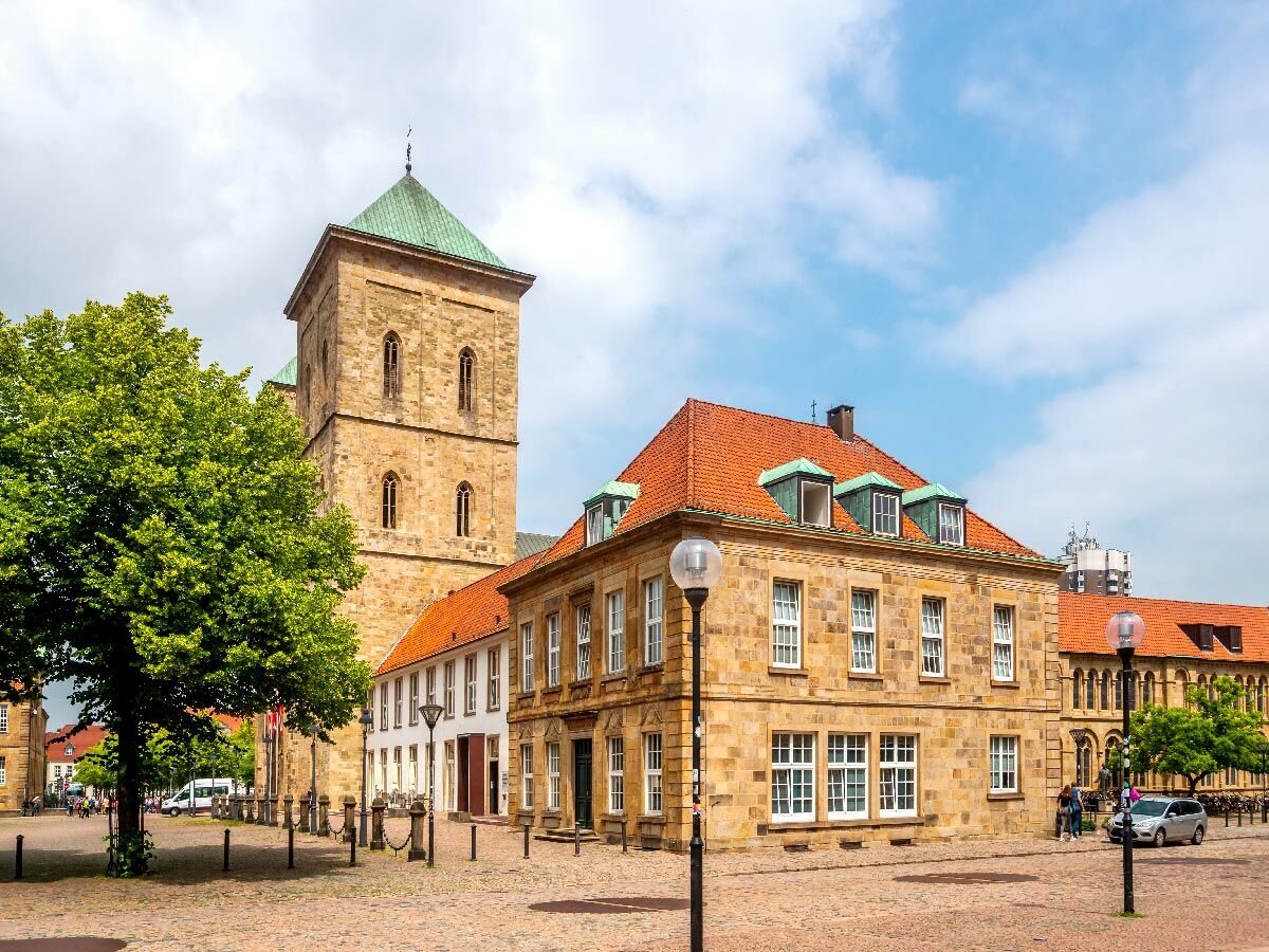 Historischer Marktplatz Paderborn mit imposantem Domturm, alten Steingebäuden, grünen Bäumen und Kopfsteinpflaster unter bewölktem Himmel.