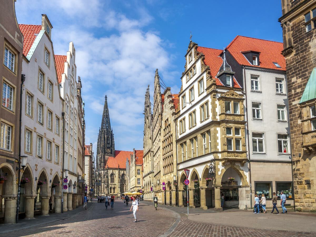Historischer Prinzipalmarkt in Münster, Deutschland, mit Giebelhäusern, Arkaden und dem Turm der Lambertikirche unter blauem Himmel.