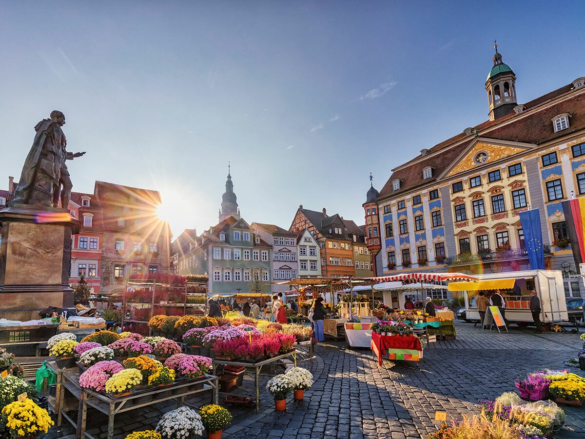Ein lebhafter Blumenmarkt auf einem sonnigen, historischen Marktplatz mit bunten Gebäuden, einer Statue und deutschen Flaggen.