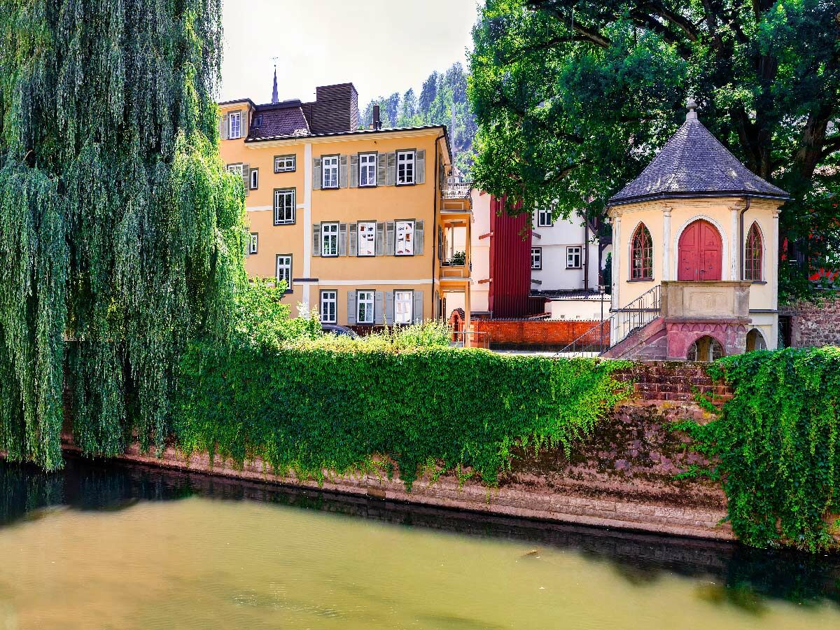 Historische Flusslandschaft mit gelbem Haus, grünem Weidenbaum und Pavillon am Wasser, umgeben von Efeu.