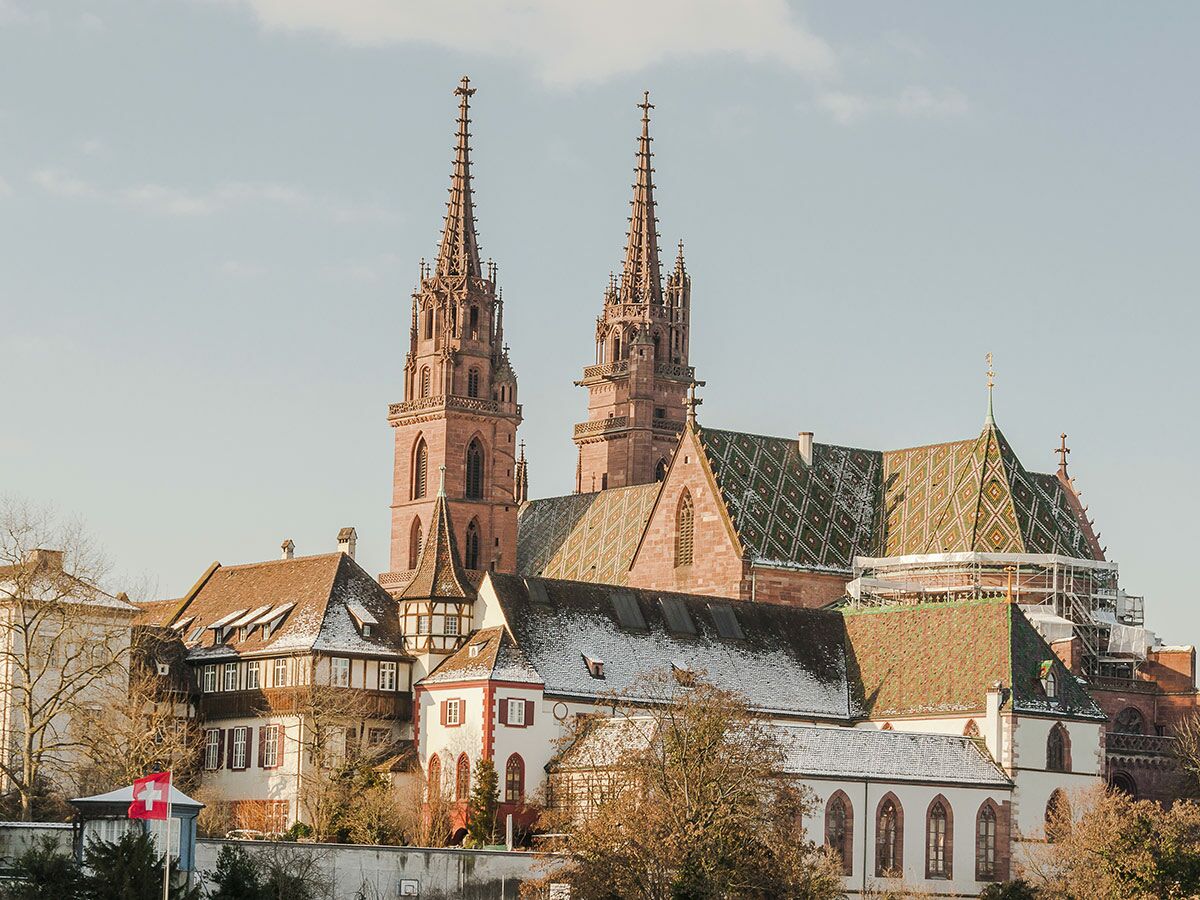 Basler Münster mit gotischen Türmen, farbigen Dachziegeln, umgeben von historischen Gebäuden mit Schnee und Schweizer Flagge.