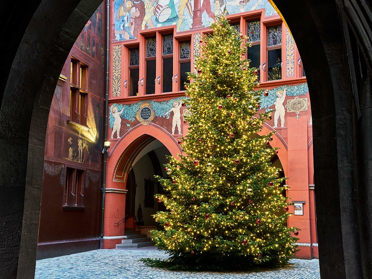 Ein festlich geschmückter Weihnachtsbaum mit Lichtern und roten Kugeln steht im Innenhof des historischen Rathauses Basel, umrahmt von Torbögen.