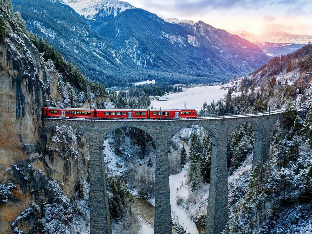 Roter Zug der Rhätischen Bahn auf dem ikonischen Landwasserviadukt in den verschneiten Schweizer Alpen bei Sonnenuntergang.