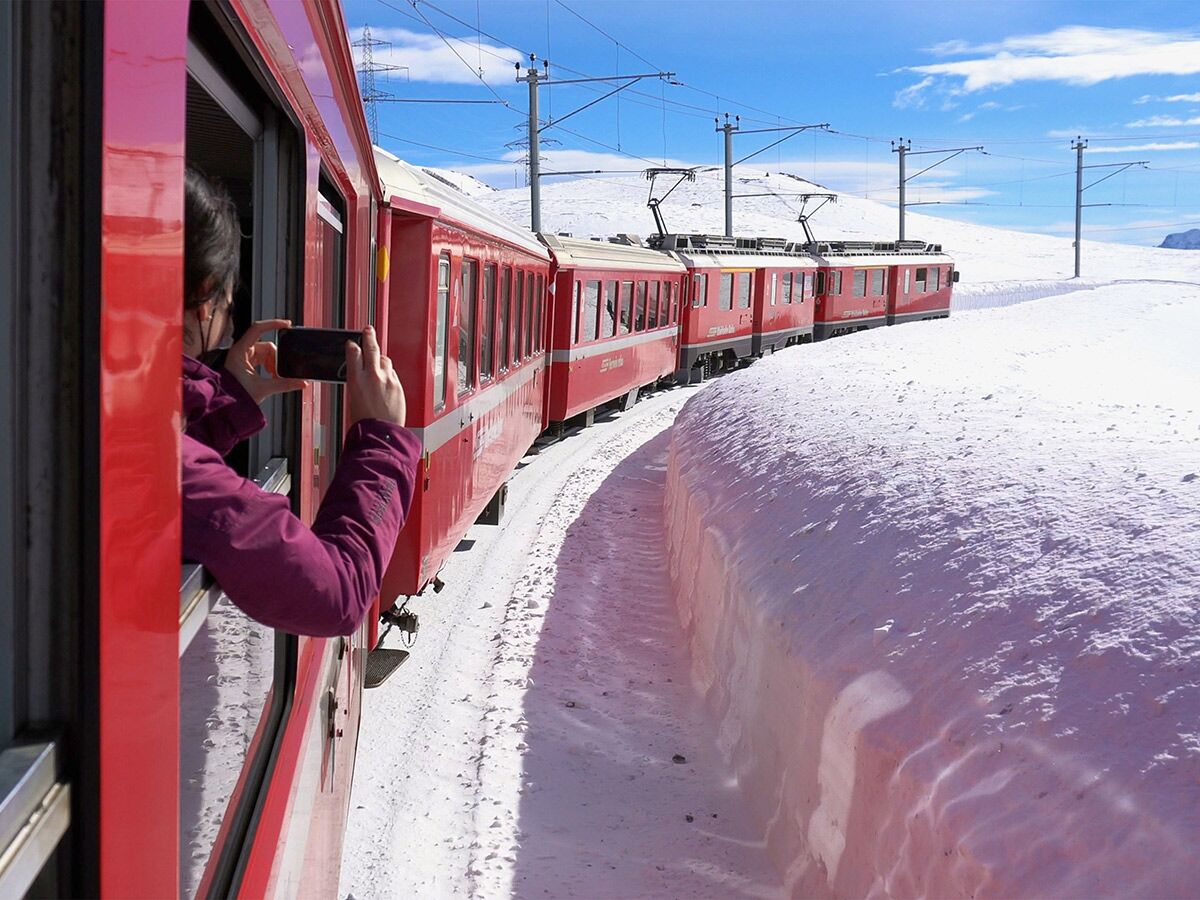 Rote Rhätische Bahn fährt durch tief verschneite Winterlandschaft. Person fotografiert mit Smartphone aus dem Fenster die Szenerie.