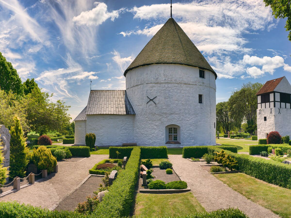 Historische weiße Rundkirche mit Kegeldach und gepflegtem Friedhof unter blauem Himmel in Dänemark.
