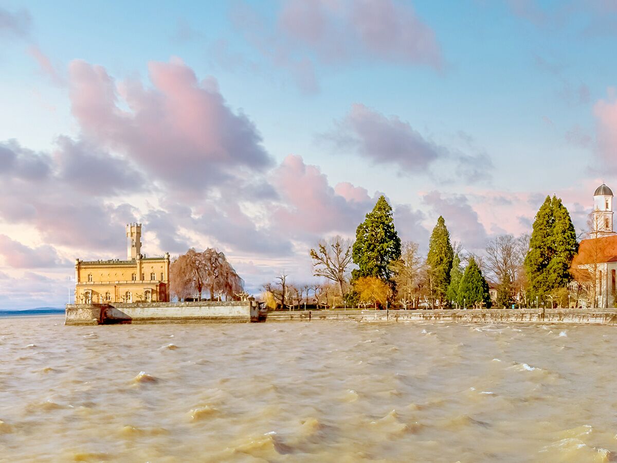 Schloss Gottlieben am Bodensee bei Sonnenuntergang. Historisches Gebäude mit Turm am Ufer, Bäume und Kirchturm im Hintergrund.