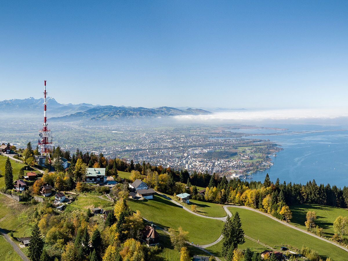 Weitblick über herbstliche Alpenlandschaft mit Bodensee, Stadt, Bergen, Häusern und rotem Sendeturm unter blauem Himmel.
