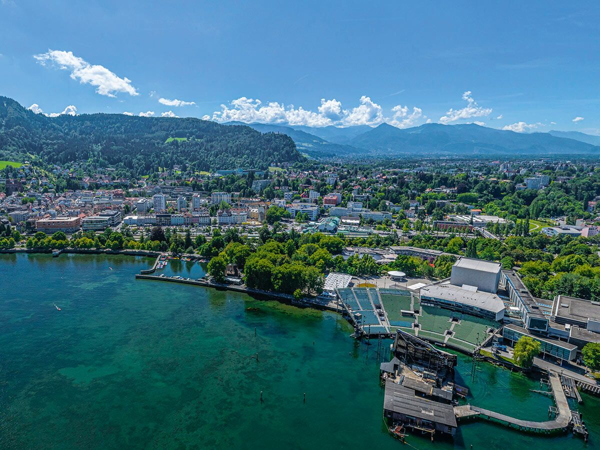 Luftaufnahme der Bregenzer Seebühne mit Stadt, Bodensee und Alpen bei strahlendem Sonnenschein.