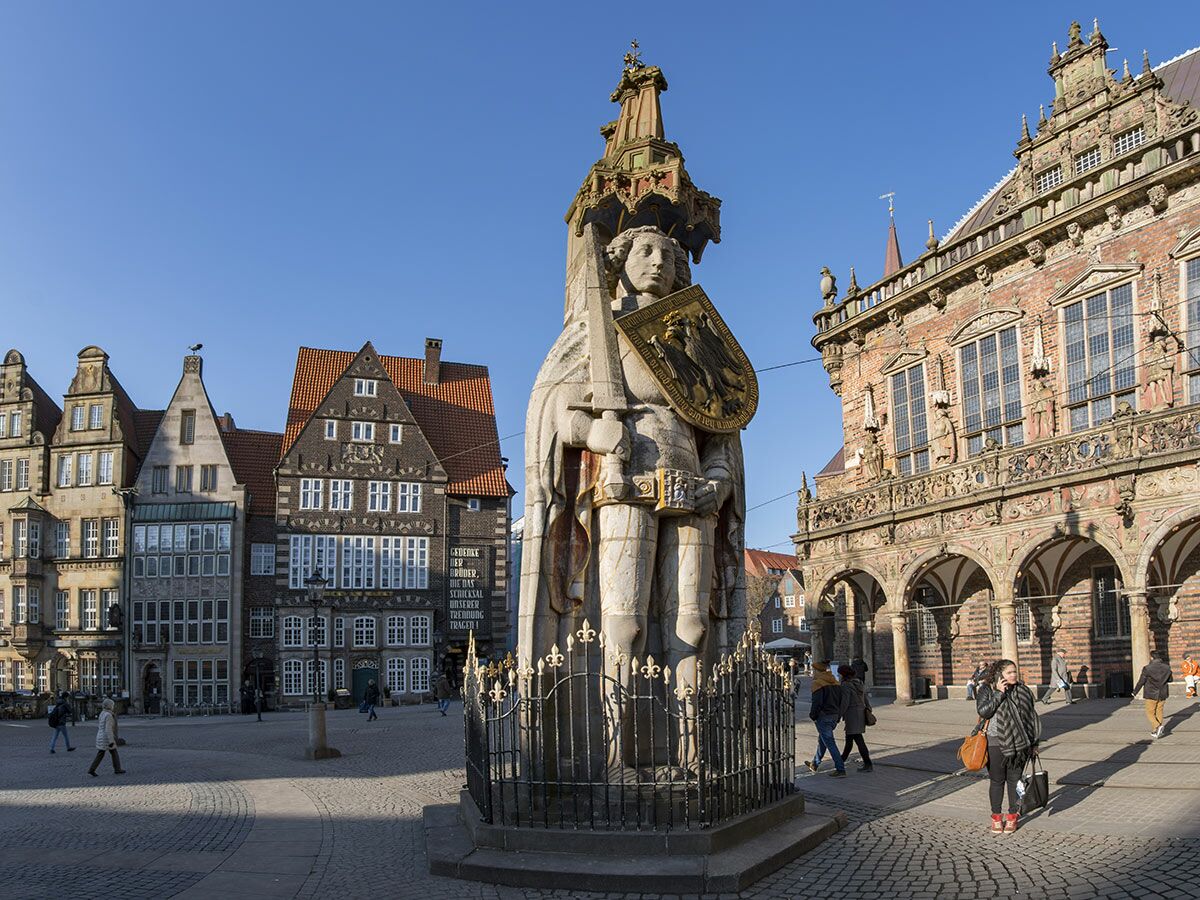 Rolandstatue auf dem Bremer Marktplatz vor dem historischen Rathaus und Giebelhäusern bei blauem Himmel, viele Passanten.