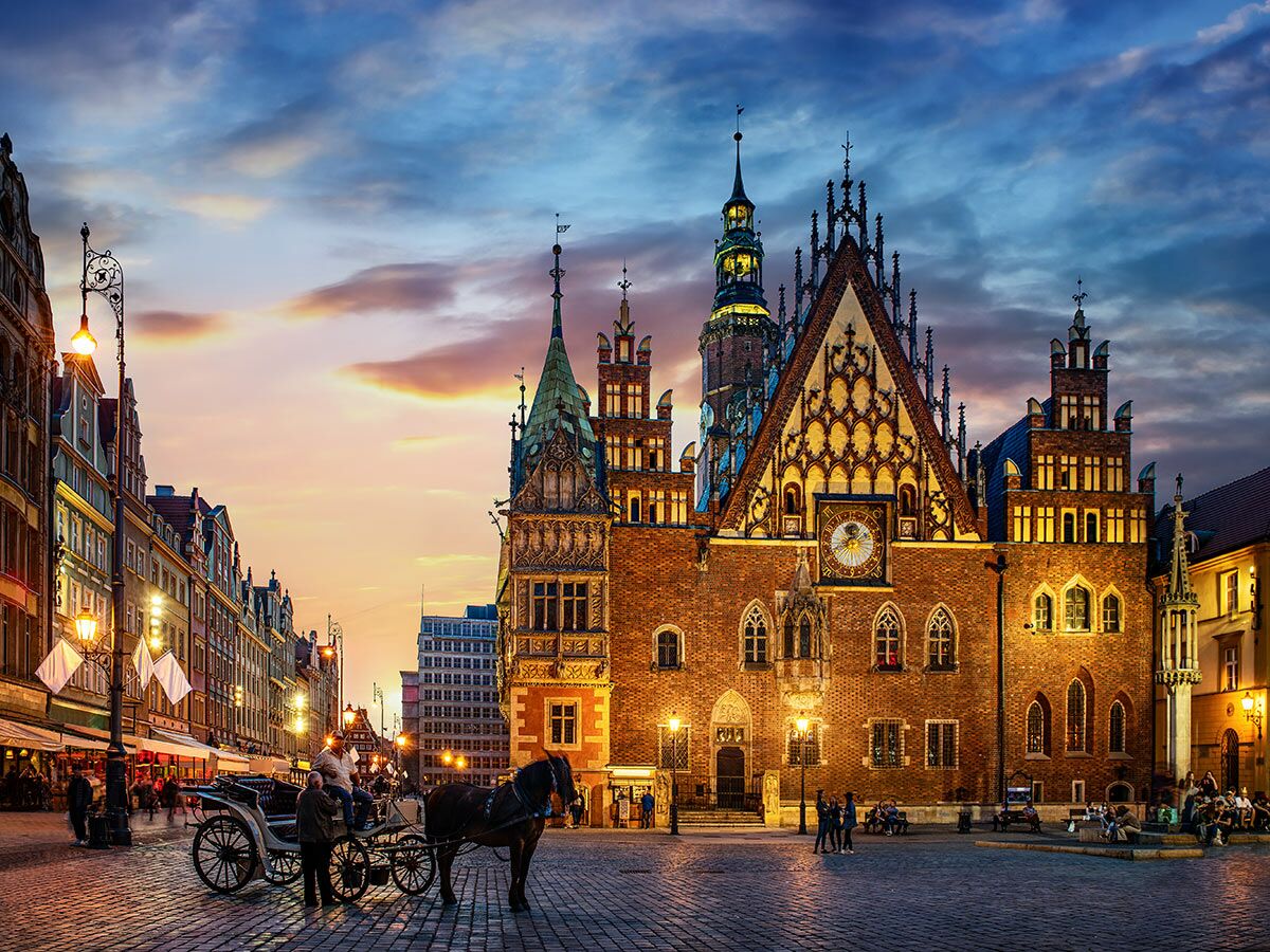 Abendstimmung auf dem historischen Altstadtplatz von Wrocław mit beleuchtetem Rathaus, Pferdekutsche und Menschen in der Dämmerung.