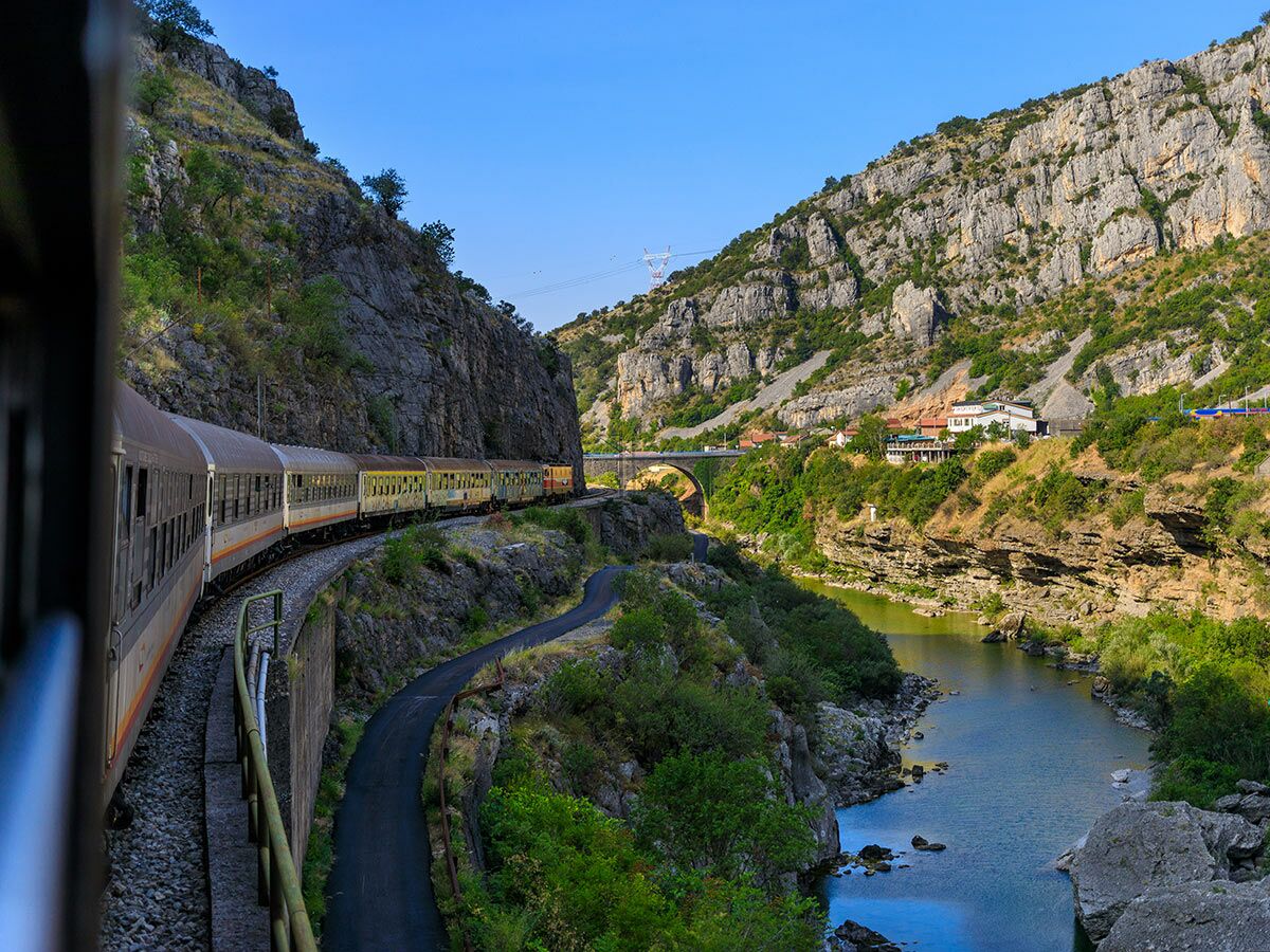 Panoramazug fährt entlang eines Flusses durch eine felsige Bergschlucht mit Brücke und Häusern unter blauem Himmel.