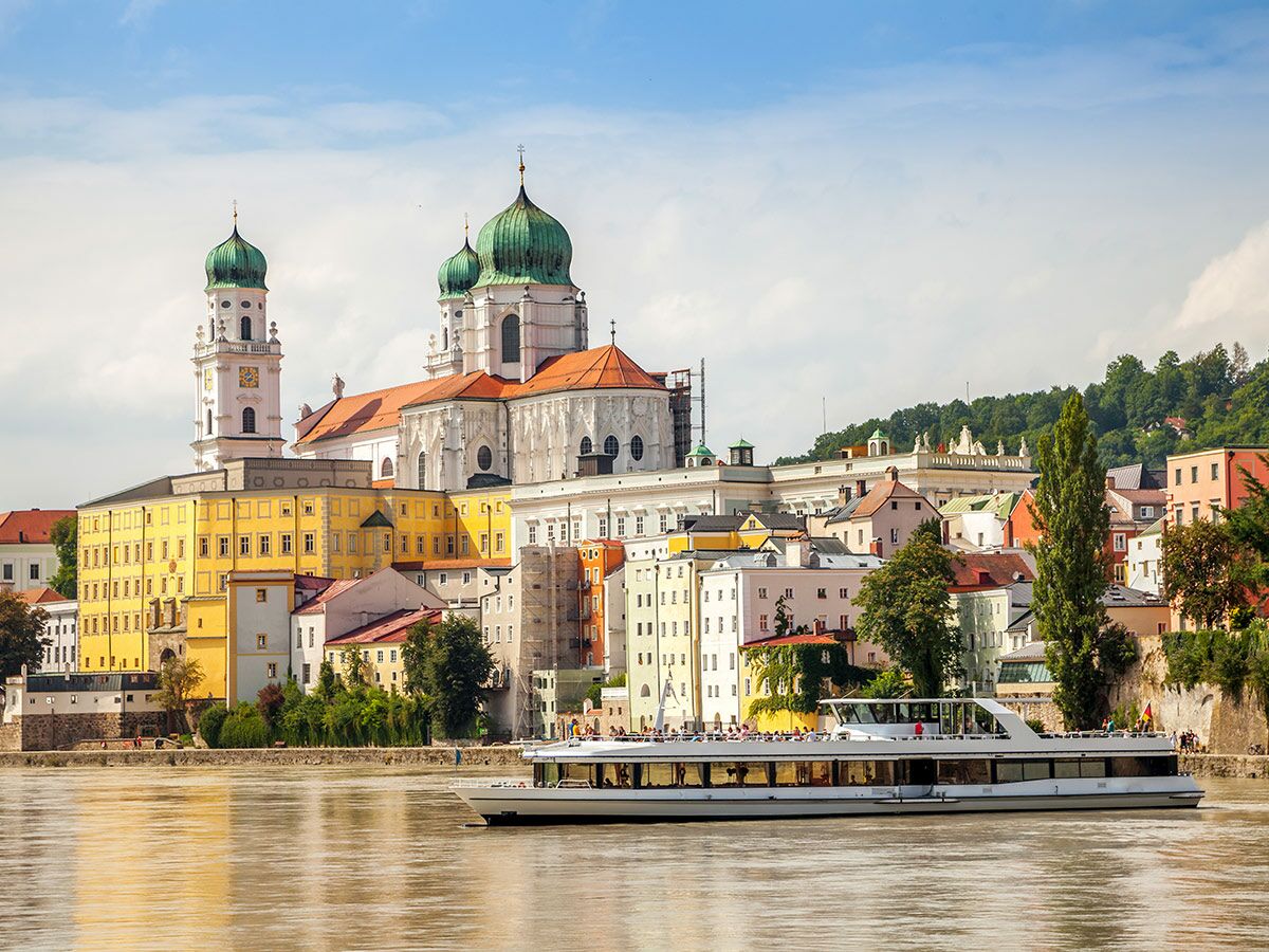 Panoramabild von Passau mit St. Stephansdom, bunten Gebäuden und einem Fahrgastschiff auf dem Fluss unter blauem Himmel.