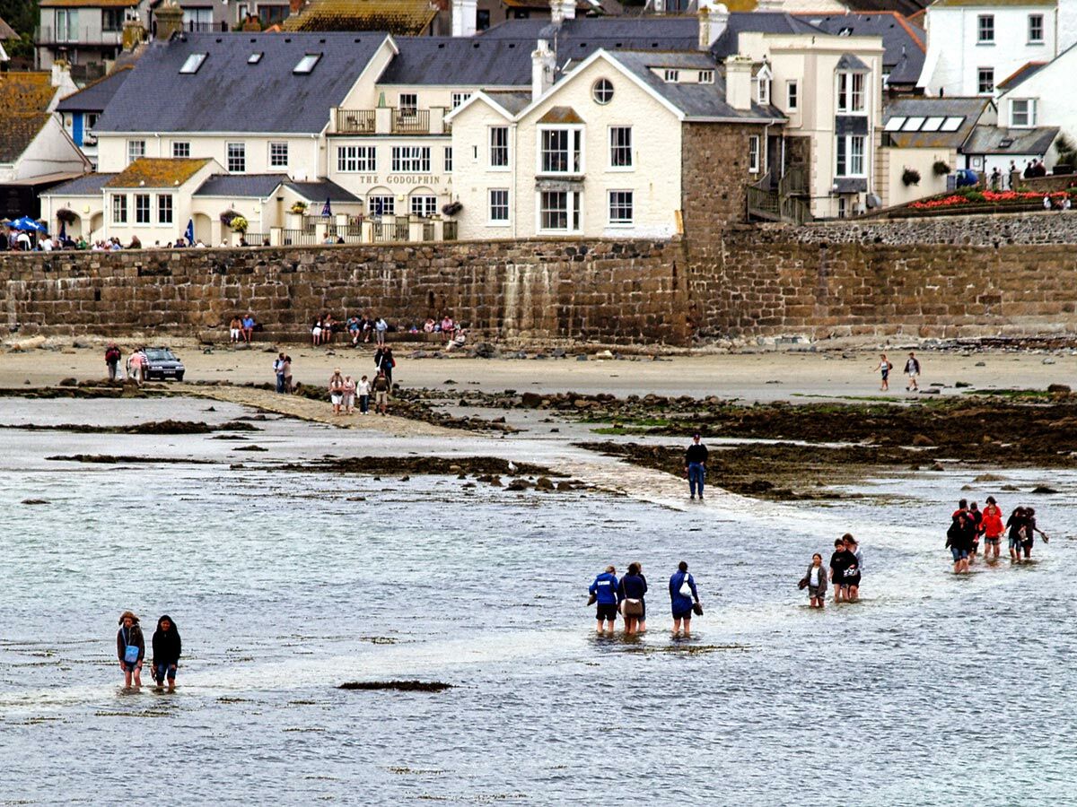 Menschen waten bei Ebbe durch das Meerwasser auf dem Weg zu St. Michael's Mount, mit Küstenstadt und Hotel im Hintergrund.