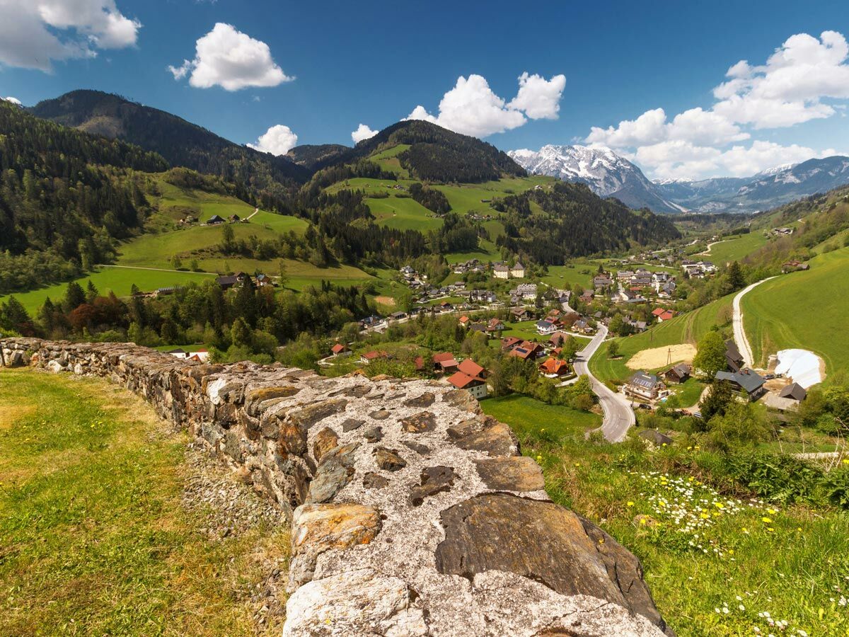 Panoramablick auf ein idyllisches Bergdorf in den Alpen, umgeben von grünen Hügeln, Wäldern und schneebedeckten Gipfeln unter blauem Himmel.