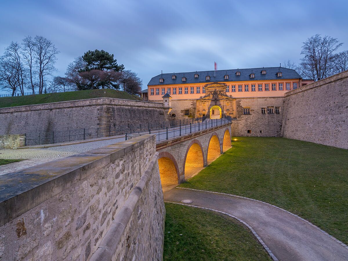 Festung Petersberg in Erfurt bei Dämmerung mit beleuchteter Bogenbrücke, beeindruckendem Tor und historischen Steinmauern.