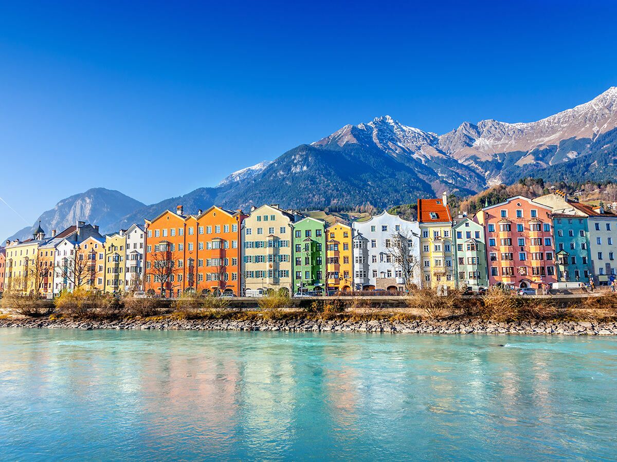 Bunte Häuserreihe am Fluss Inn in Innsbruck, dahinter schneebedeckte Alpen unter blauem Himmel.