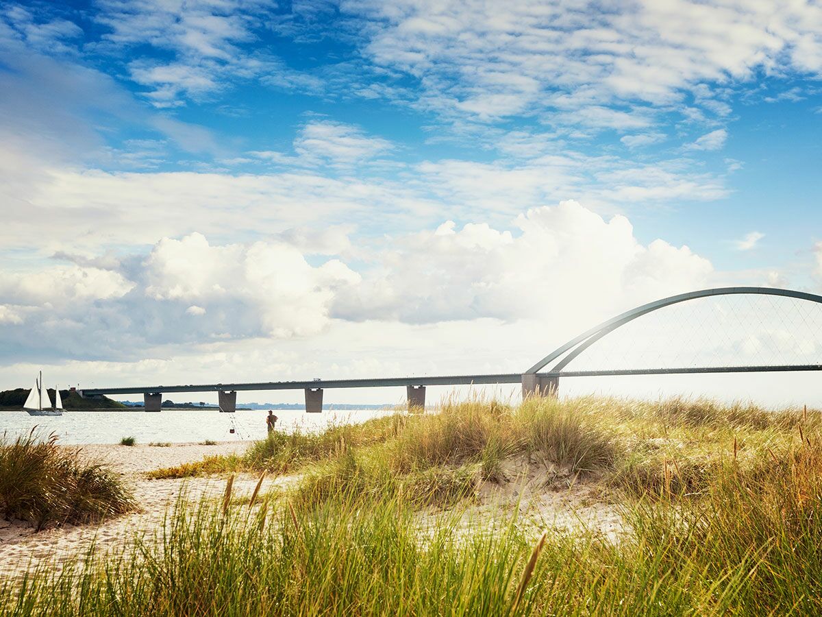 Panoramablick auf eine moderne Bogenbrücke über das Meer, dahinter eine Küstenlandschaft mit Sandstrand, Dünen und einer Person am Ufer.