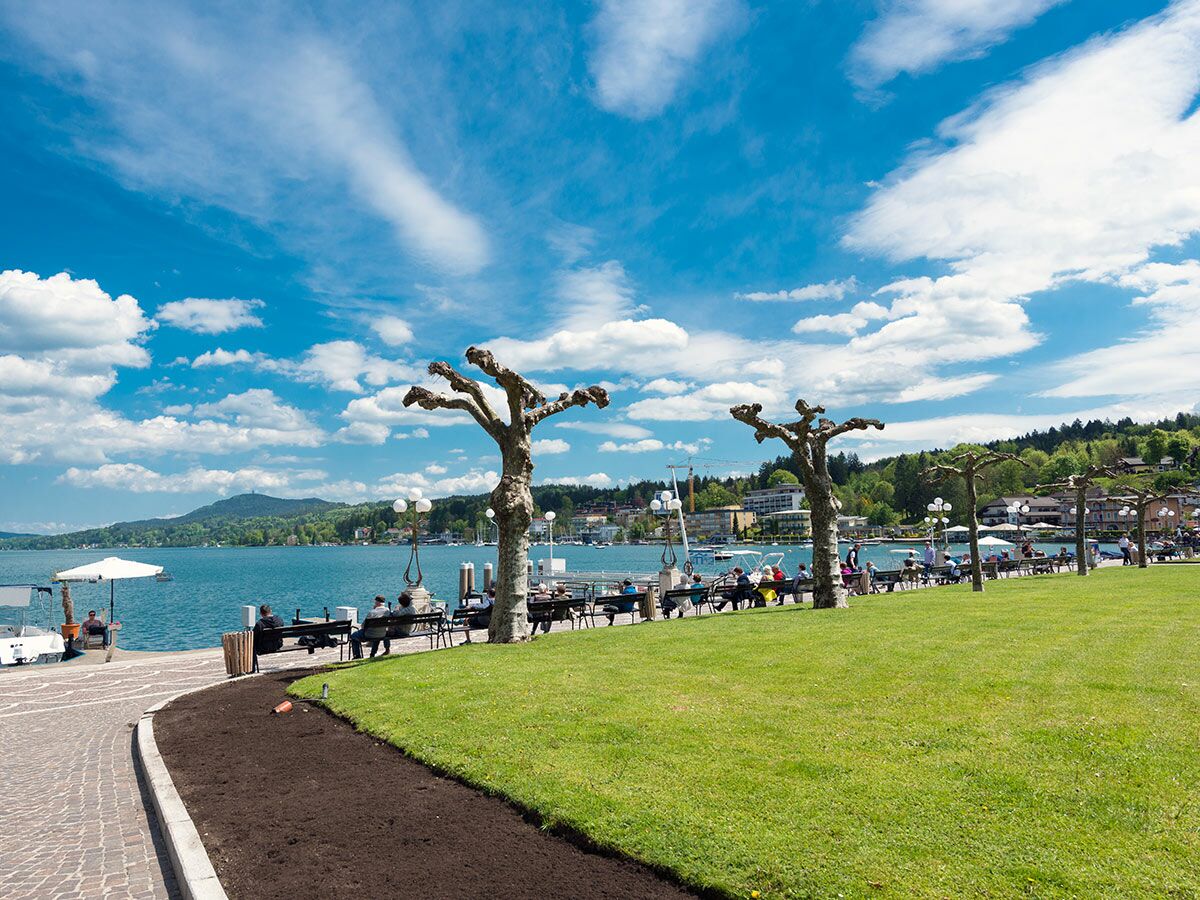 Menschen entspannen auf Bänken entlang der grünen Uferpromenade des Wörthersees unter blauem Himmel mit Wolken.