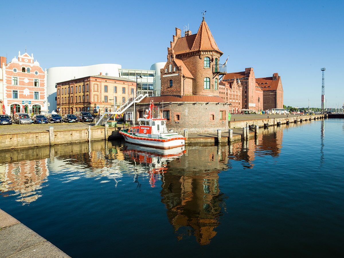 Panorama des Rostocker Hafens mit historischen Backsteingebäuden, Boot im Wasser, klarem Himmel und Spiegelungen.