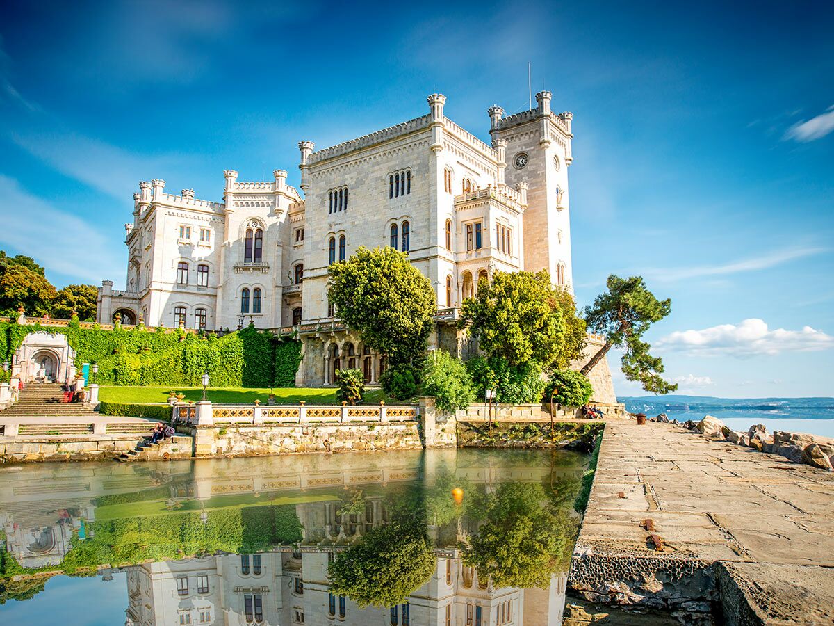 Weißes Schloss Miramare an der Adria, mit Gärten und Reflexionen im Wasser bei blauem Himmel.