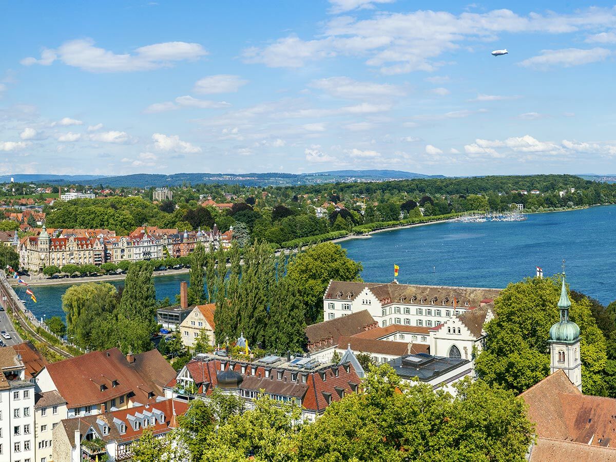 Panoramablick auf Konstanz am Bodensee mit Altstadt, Seeufer, Jachthafen, grünen Bäumen und einem Zeppelin am Himmel.