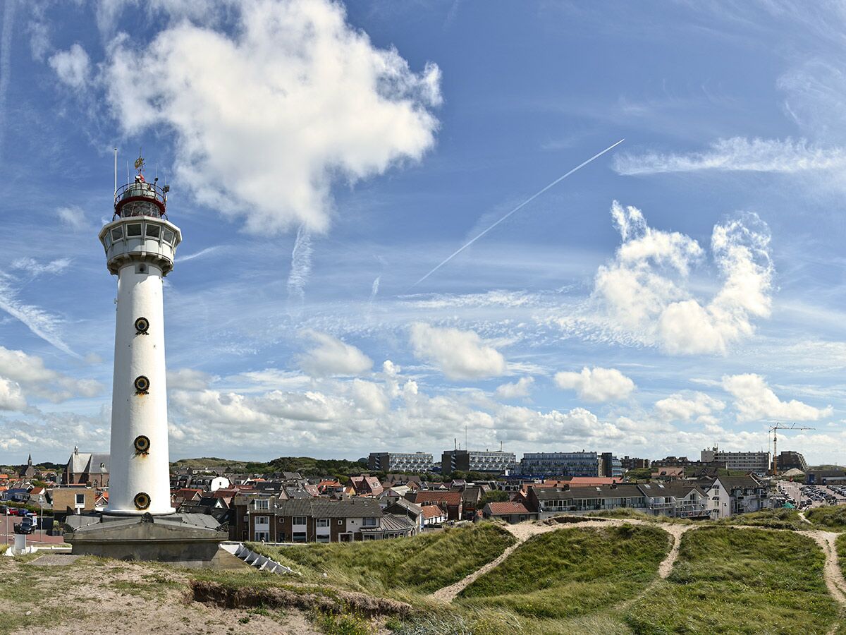 Weißer Leuchtturm in einer Küstenstadt mit Häusern, Dünen und blauem Himmel. Panoramaaufnahme bei Tag.