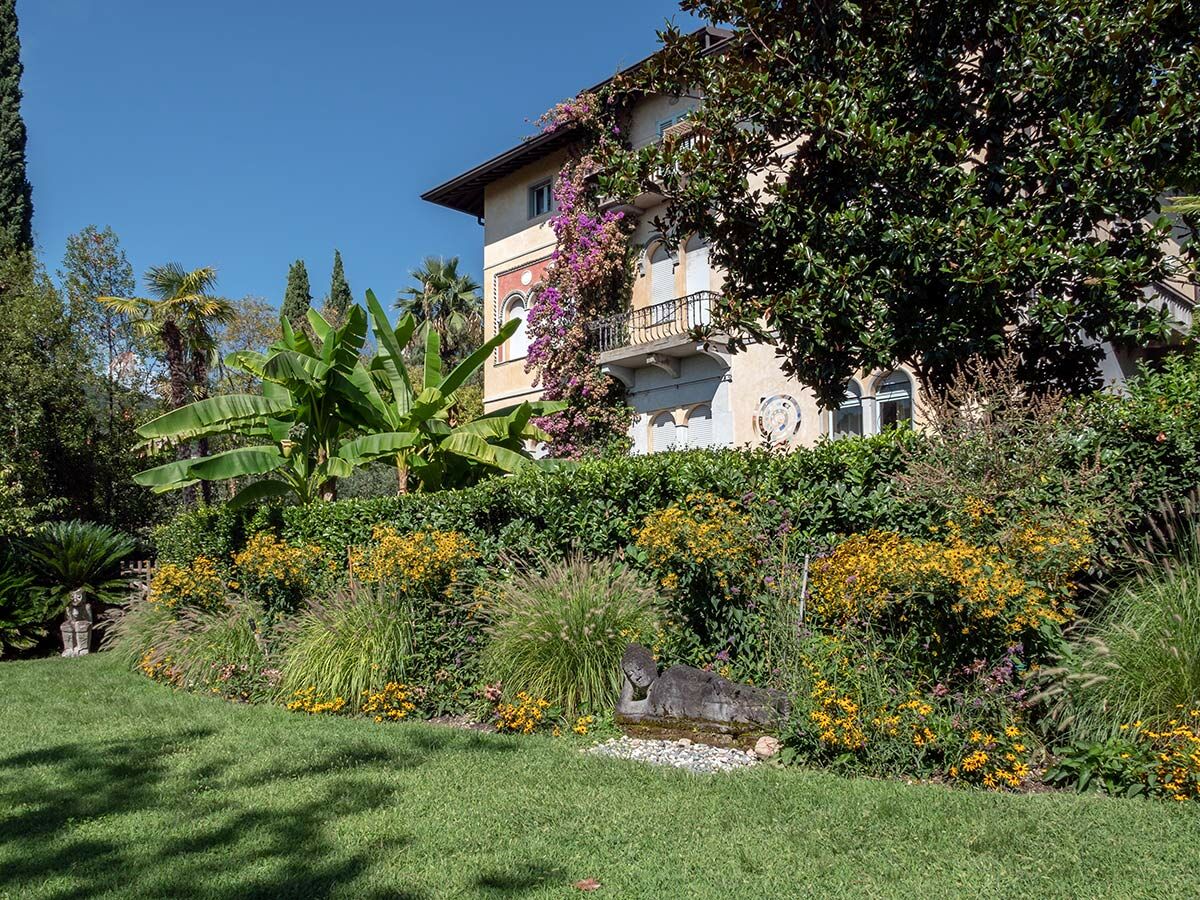Klassische Villa mit mediterranem Garten, üppigem Rasen, bunten Blumen, Palmen und Bananenstauden unter blauem Himmel.